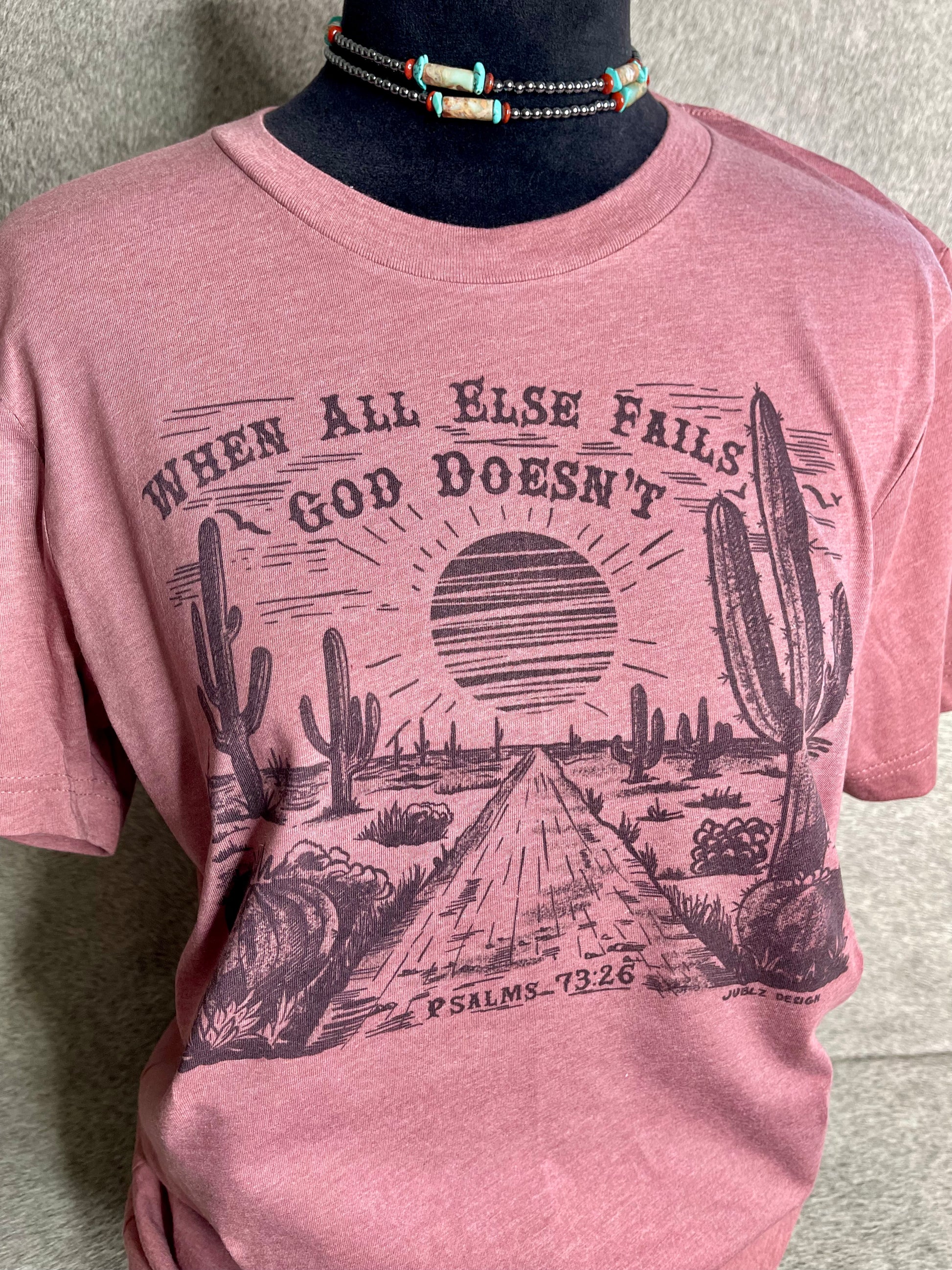 Soft heather mauve Bella + Canvas unisex t-shirt featuring a sublimation desert scene with the faith-inspired quote "When All Else Fails, God Doesn’t" at the top and "Psalms 73:26" at the bottom. A stylish and meaningful Christian apparel piece with a Western touch.