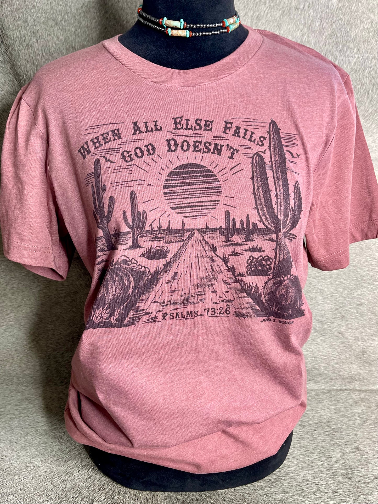 Soft heather mauve Bella + Canvas unisex t-shirt featuring a sublimation desert scene with the faith-inspired quote "When All Else Fails, God Doesn’t" at the top and "Psalms 73:26" at the bottom. A stylish and meaningful Christian apparel piece with a Western touch.