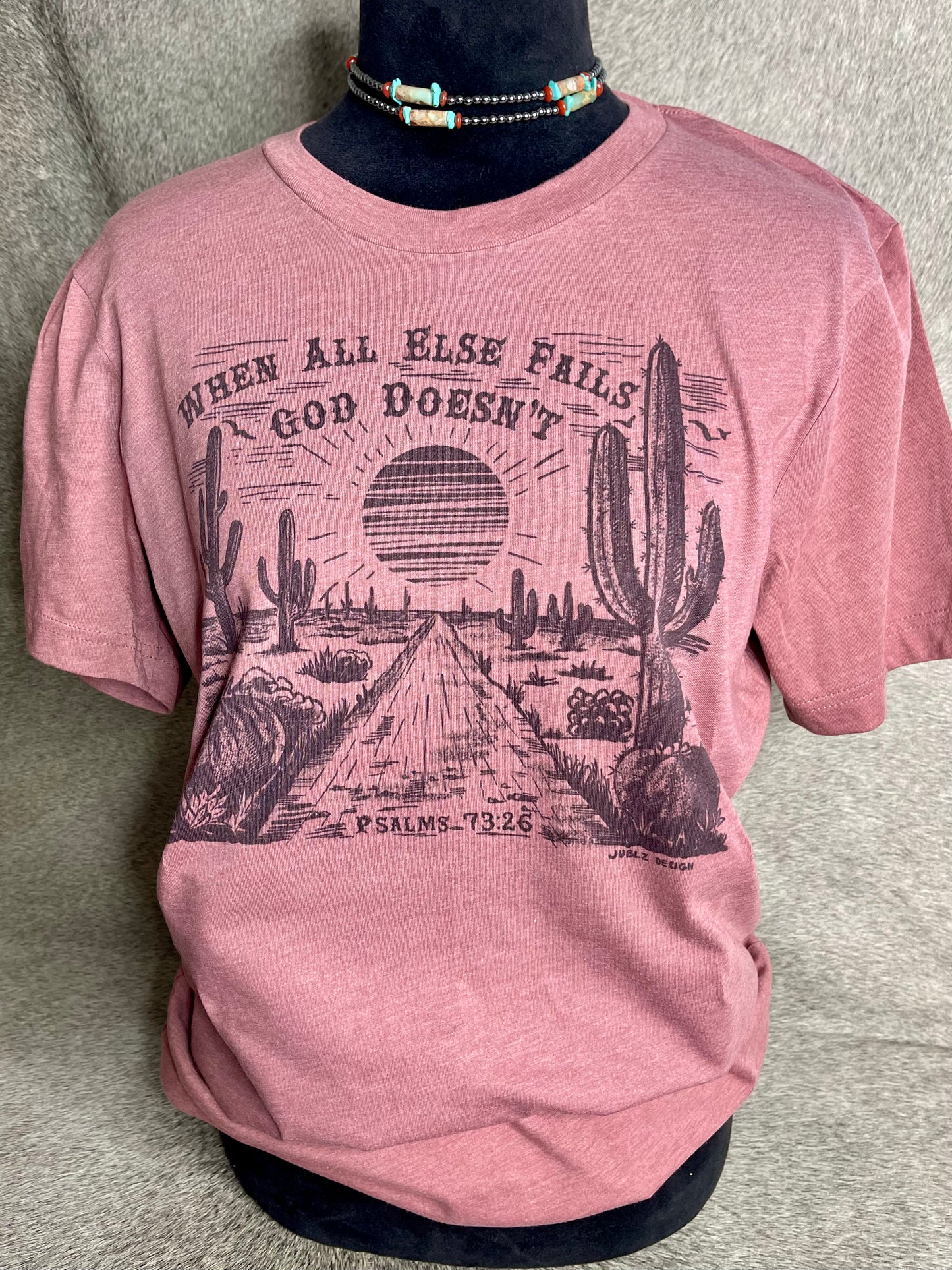 Soft heather mauve Bella + Canvas unisex t-shirt featuring a sublimation desert scene with the faith-inspired quote "When All Else Fails, God Doesn’t" at the top and "Psalms 73:26" at the bottom. A stylish and meaningful Christian apparel piece with a Western touch.
