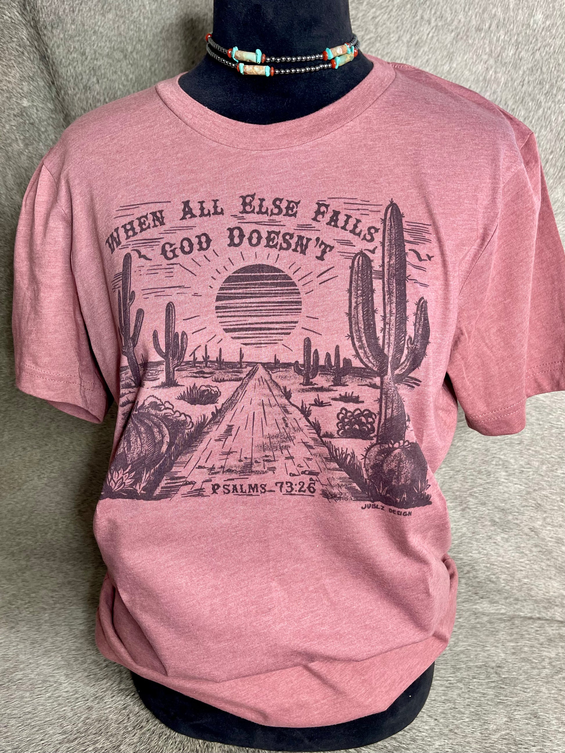 Soft heather mauve Bella + Canvas unisex t-shirt featuring a sublimation desert scene with the faith-inspired quote "When All Else Fails, God Doesn’t" at the top and "Psalms 73:26" at the bottom. A stylish and meaningful Christian apparel piece with a Western touch.