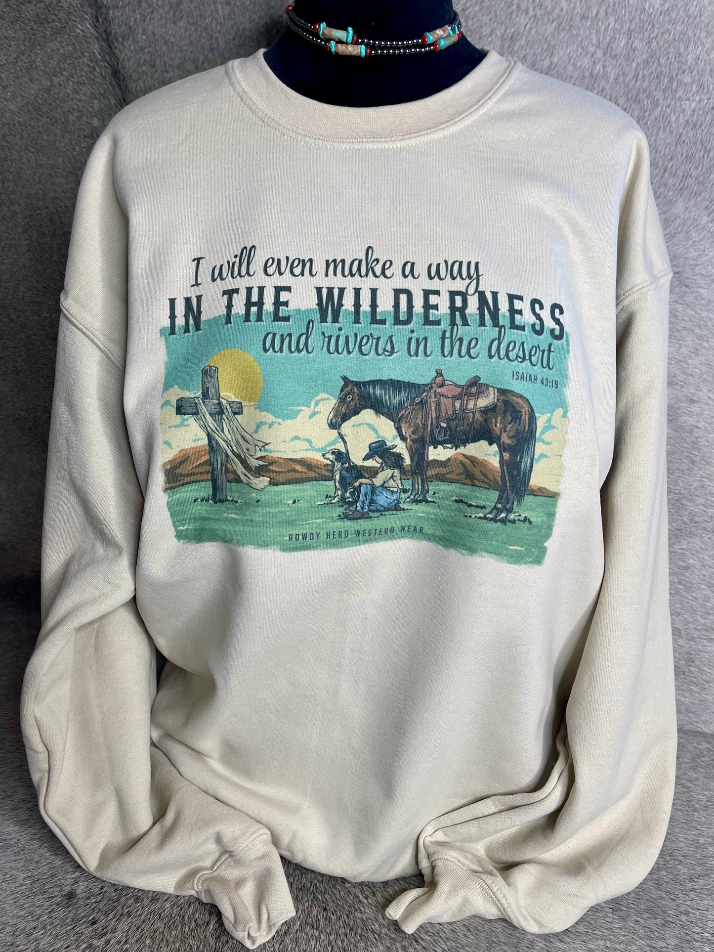 A unisex Gildan sweatshirt in sand color featuring a sublimated design of a praying cowgirl with her dog and horse, along with the Isaiah 43:19 quote.