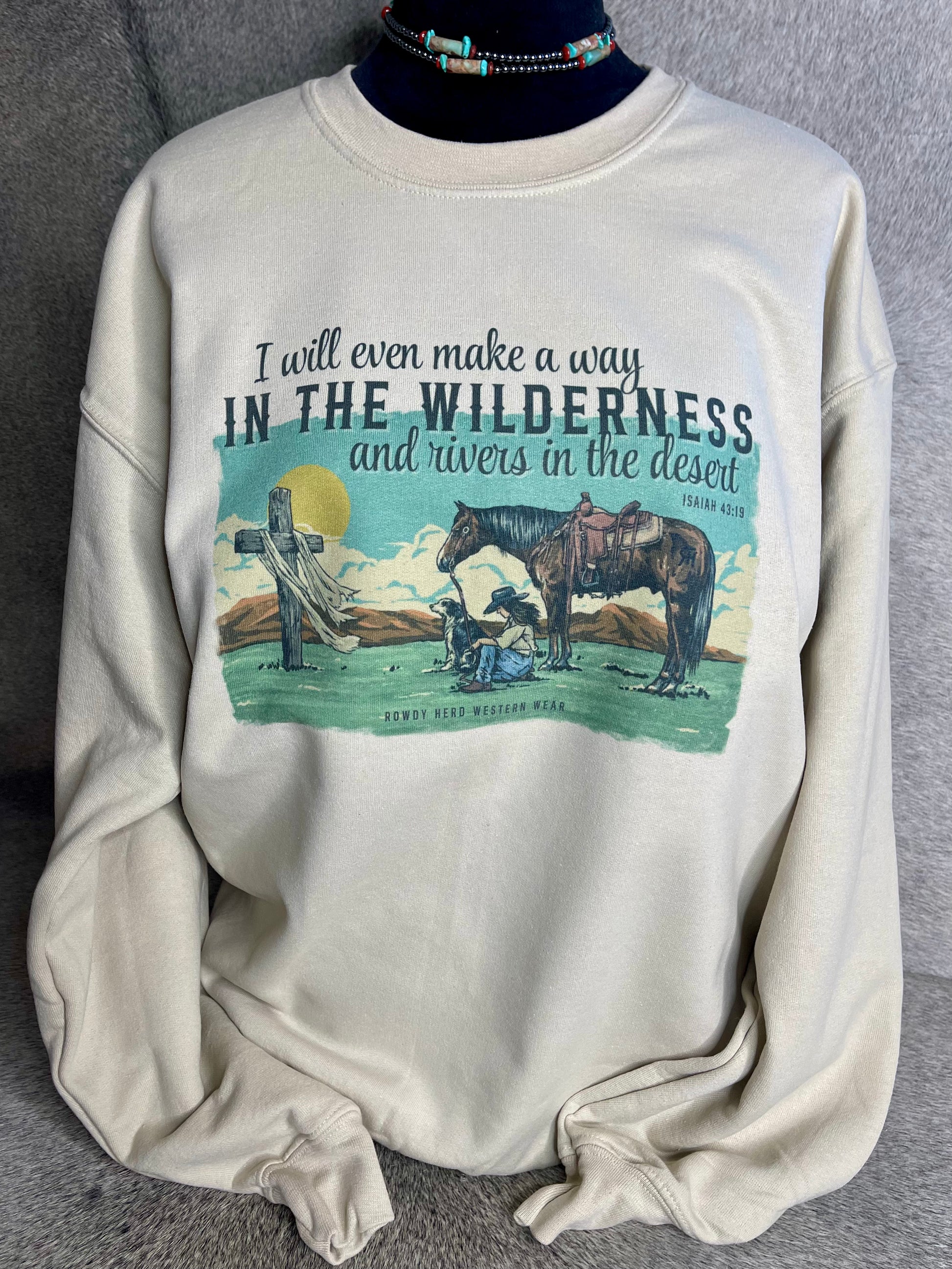 A unisex Gildan sweatshirt in sand color featuring a sublimated design of a praying cowgirl with her dog and horse, along with the Isaiah 43:19 quote.
