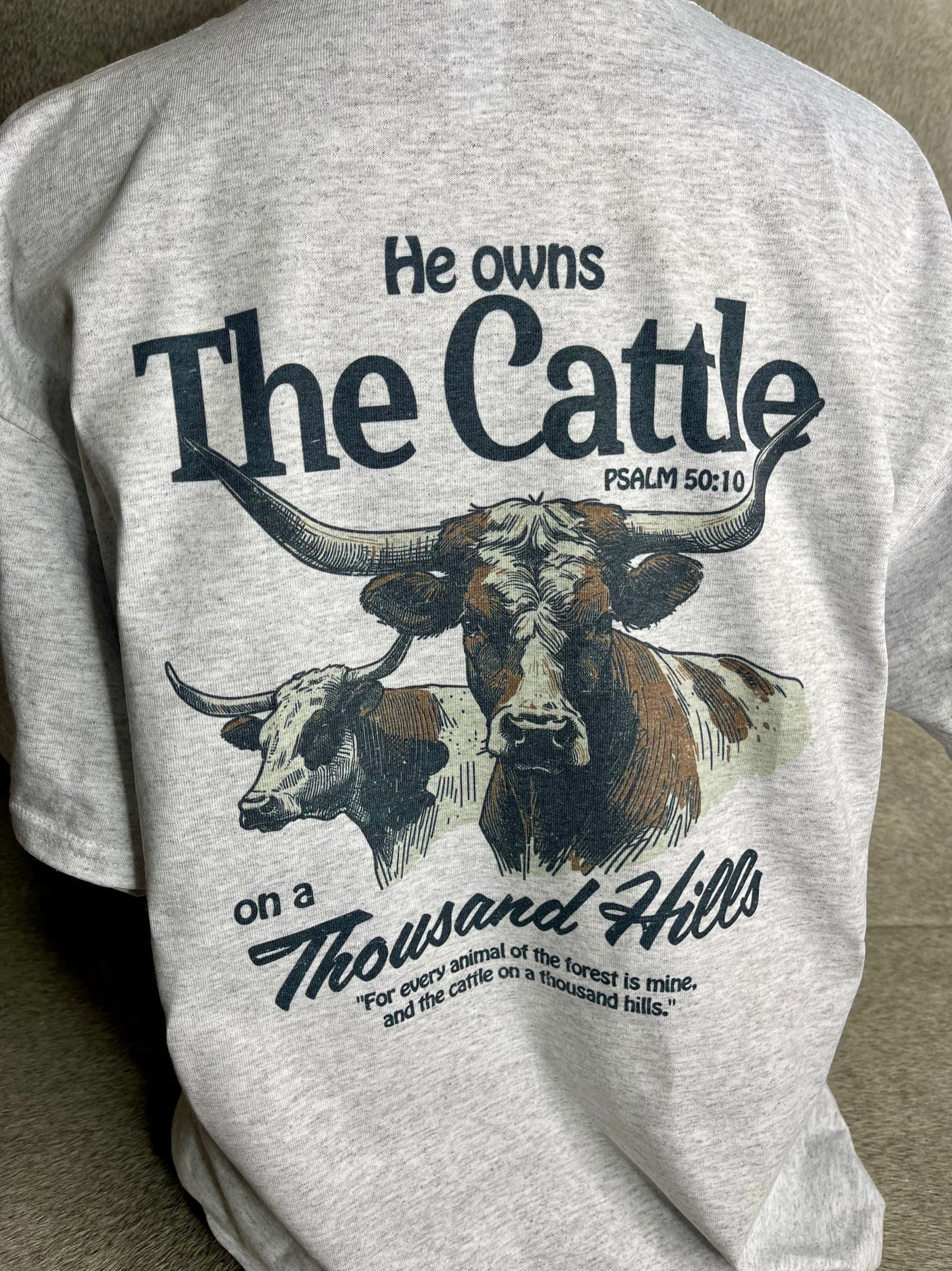 A heather grey Jerzees unisex t-shirt featuring the Rowdy Herd logo on the front pocket area and a longhorn cattle design on the back, along with the quote from Psalms 50:10.