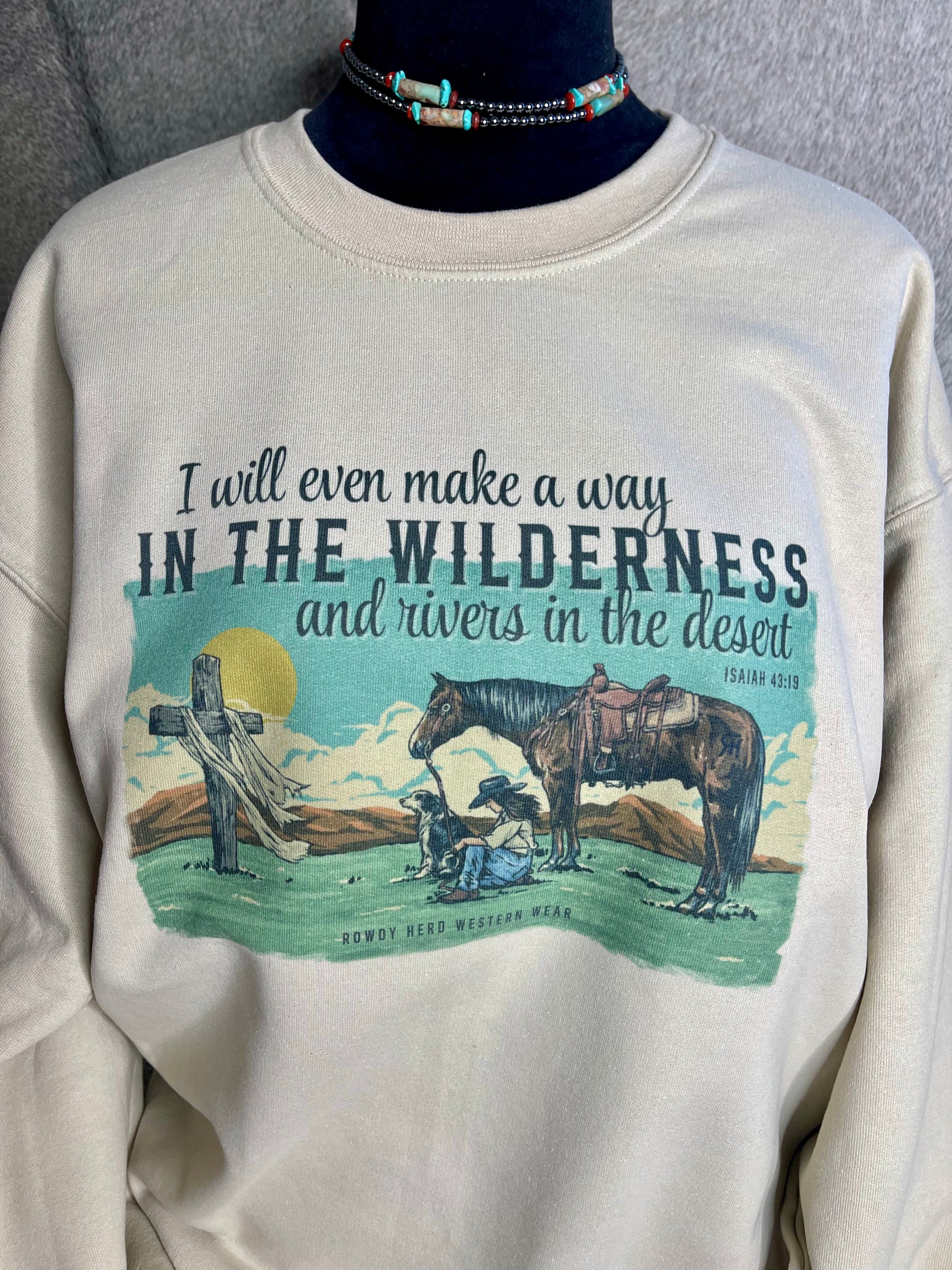 Gildan unisex sweatshirt in sand, featuring a stunning design of a praying cowgirl surrounded by her dog and horse, with Isaiah 43:19 elegantly quoted